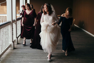 A bride in a white dress walks happily down a wooden hallway, holding her gown, followed by bridesmaids in dark dresses. The scene is joyful and candid, with natural light streaming in from the left.