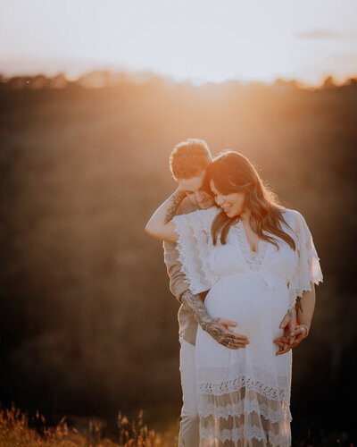 Pregnant lady with man hugging her from behind, his hands on her belly. Golden sunlight spills from the top of the frame.