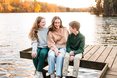 3 siblings sit laughing on a dock at Peters Lake Park in Canonsburg during their fall family photography session.