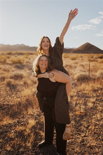 Andi gives Steph a piggyback ride in a sunny desert setting. Steph joyfully raises an arm to the sky, while both smile brightly against a backdrop of dry grass and distant mountains.