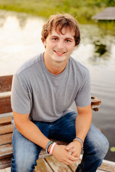 A young man with short brown hair sits on a wooden bench by a body of water, captured beautifully by Amber Bauer Photography. He is wearing a gray t-shirt and blue jeans, smiling at the camera. The serene Brainerd Lakes backdrop features calm water and lush green grass.