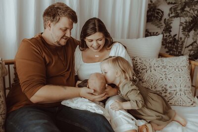 A family of four sits on a couch in a Macon studio. A man holds a baby while a woman smiles, capturing the essence of newborn joy. A small child leans in to kiss the baby, surrounded by neutral tones and decorative pillows.