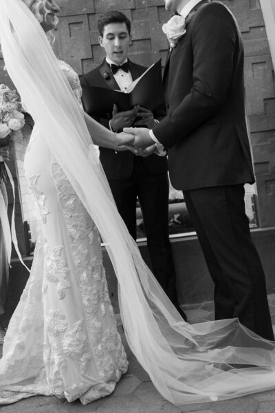 A bride and groom holding hands during the ceremony and veil flowing through the wind.