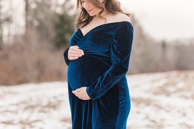 Woman stands in snowy field  during winter maternity photo session with Sara Sniderman Photography in Natick  Massachusetts