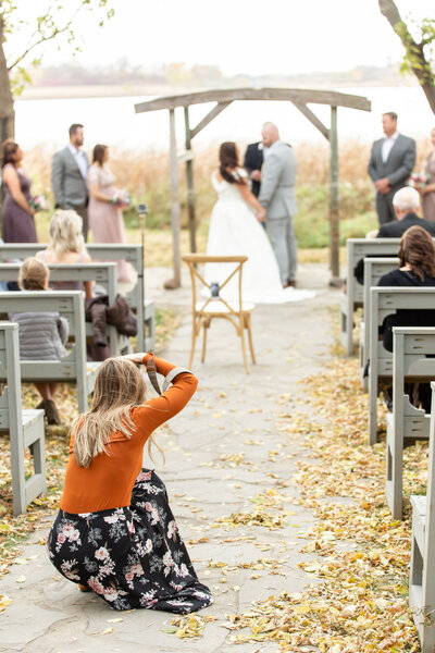 Abby is crouched down in the aisle, capturing an outdoor wedding ceremony