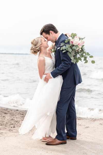 Bride and groom stand closely nose to nose smiling at each other on the beach