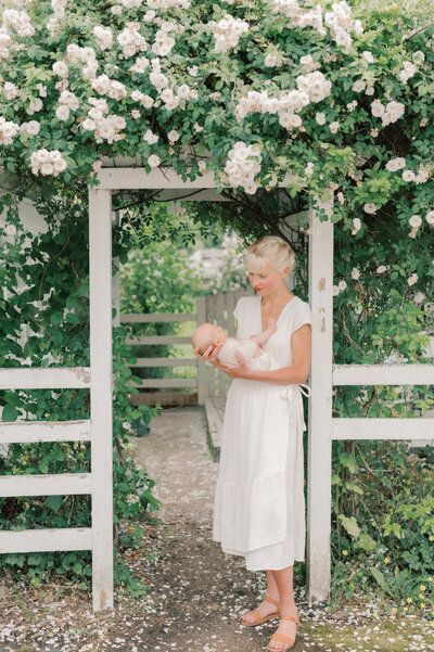 Mother standing under a white rose arbor holding her newborn baby