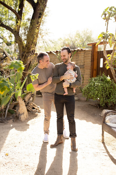 Two dads smiling and holding their baby during a warm, sunlit family session outdoors, captured by a Palm Springs family photographer.