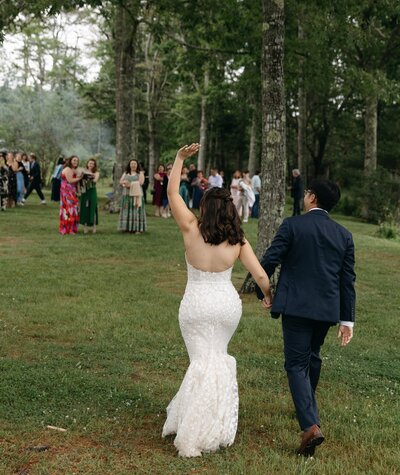 New England couple holding hands with the bride waving to guests, captured from behind at their wedding."