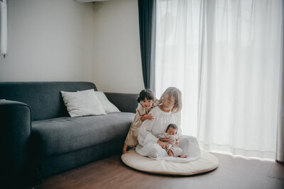 窓際での親子写真。新生児を膝に乗せた優しい笑顔のお母さんにくっつく子ども。