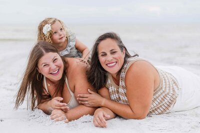 Two moms laying in the sand with their toddler daughter climbing on their back