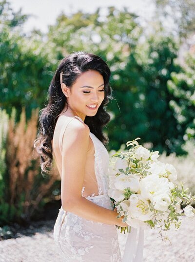 Kohnur bride with full glam makeup and long Hollywood waves with a side clip, holding a white bouquet in California.