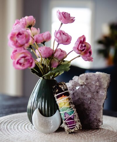a vase of pink flowers next to a purple crystal, burnt sage, and a crystal shaped like a crescent moon