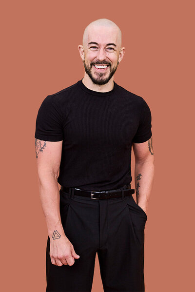 Jake, Male hairstylist with a shaved head, wearing a fitted black shirt and trousers, showcasing tattoos on arms.