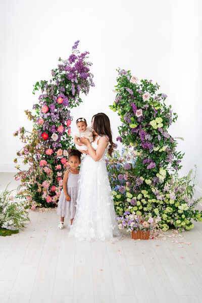 A mother in a flowing white gown holds a smiling baby while a young girl in a lavender dress stands close by, all framed by vibrant floral arches in a light-filled studio.