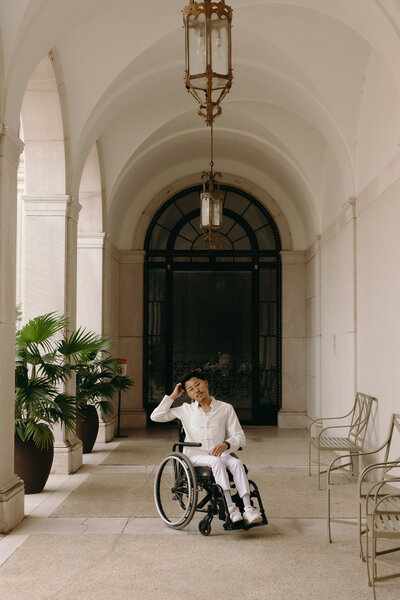 A woman wearing a scarf around her head with big sunglasses, whit gloves, and a yellow blazer over a jean vest with a long jean skirt. Her brown bag is swinging in front of her with one hand on her head
