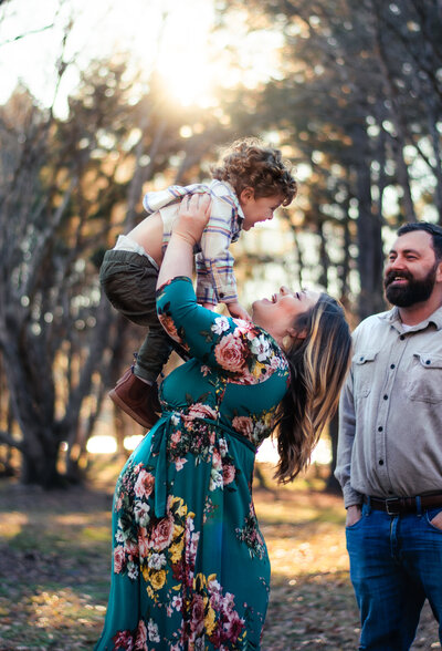 Pregnant mom in floral dress lifts young toddler with curly hair in the air as dad watches them smiling and in love with his family