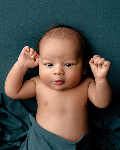 Awake newborn looking at the camera, lying on a teal background.