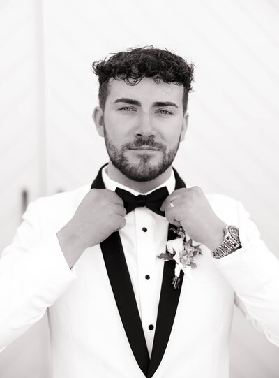 A bride in a white gown and veil holds pink flowers, facing a smiling groom in a dark suit. They are outdoors on a sunny day, with trees in the background.