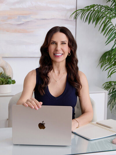 ennifer Taormina, founder of Jennifer Taormina & Co., sitting at her desk with a MacBook, notebook, coffee mug, and plant décor in a bright, modern office.