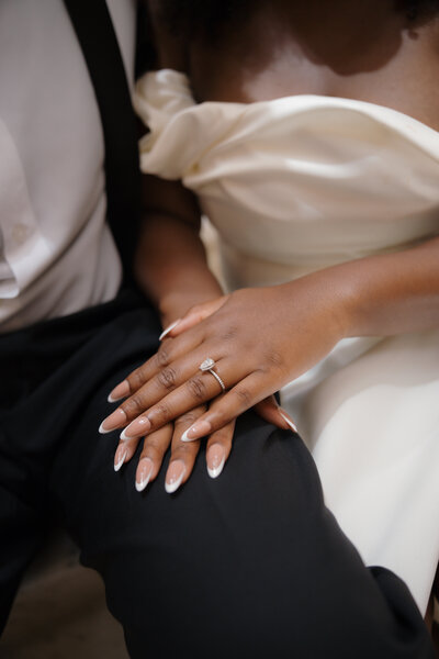 Close-up of a bride’s hand resting on her groom’s leg, showcasing her engagement ring and elegant nails.