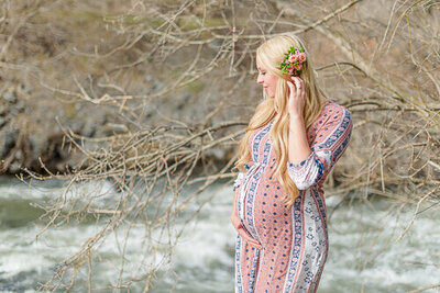 A pregnant woman wearing a pink maternity dress stands by a river in Provo Canyon. Captured by Utah maternity photographer Melissa Woodruff Photography