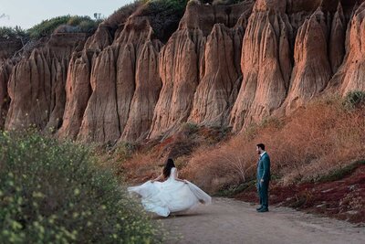 A bride in a full, layered princess gown spins gleefully as she and her groom walk down a sandy dirt path to access a private beach during their luxury adventure elopement in California.  behind them are striking red dirt cliffs, with hints of yellow wildflowers in the frame.  