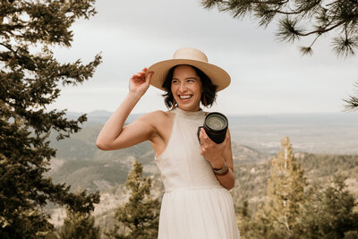 colorado elopement photographer touches hat and poses with her camera in the colorado mountains.