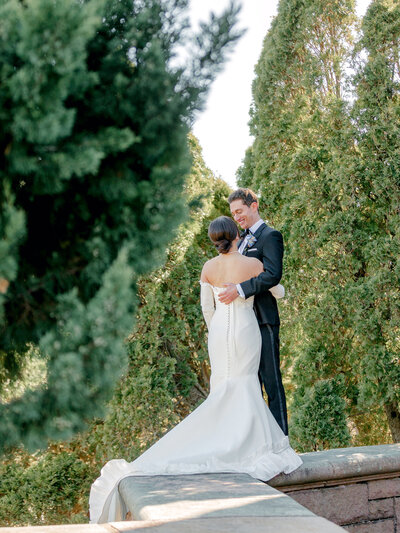 Bride in a white gowns with removable sleeves and groom in a black tux standing on a retaining wall, smiling at each other, with pine in the foreground and background