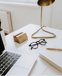 Partial view of a white desk. An open MacBook, pencil holder, black-rimmed glasses, a white book and gold pen, and a brass desk lap are displayed on a white desk. In the background is a white wall and white radiator.