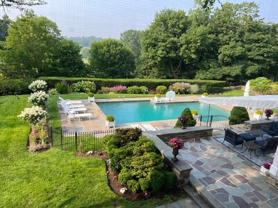 A beautifully landscaped backyard featuring a luxurious pool with stone decking, lush greenery, flowering plants, and an outdoor seating area, creating an inviting and elegant outdoor space.