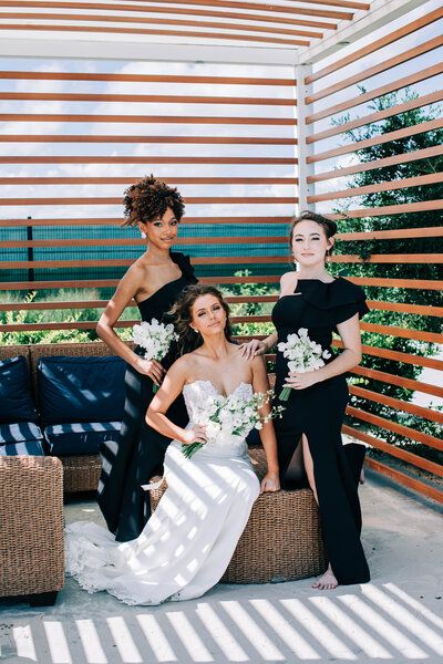 Bride in a strapless gown with two bridesmaids in black dresses posing under a slatted outdoor pergola lounge.