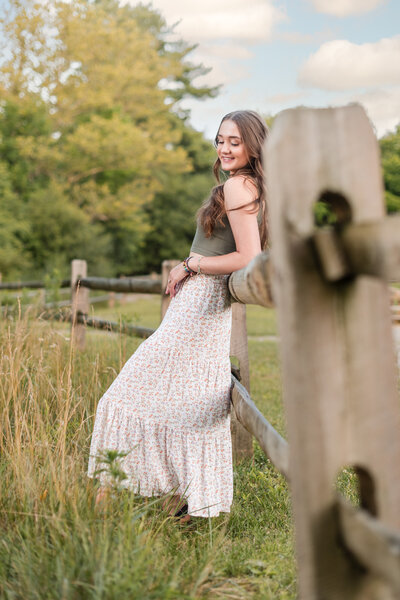 Sun-filled image of high school senior girl in off-white off the shoulder sweater leaning on wooden fence