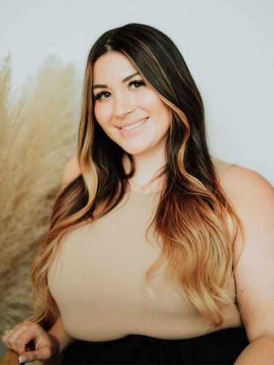 A woman with shoulder-length dark hair, wearing a black blazer, sits on a brown chair and smiles at the camera. Beige pampas grass is visible in the background, reflecting the modern ambiance of Falmouth Maine Medical Aesthetics.