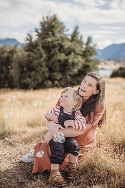 mum and son sitting in the grass watching bubbles during a family photography session in cromwell