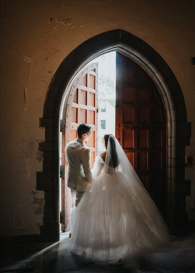 Bride and Groom looking out the gorgeous arches door from inside the church, while holding hands. The bride has a veil on and the groom has an ivory suit