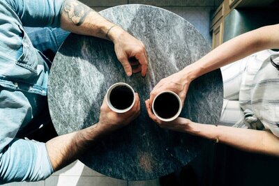 Overhead view of two people sitting at a round stone table, each holding a cup of coffee in a casual conversation setting.