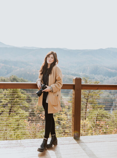 Woman in black outfit with tan cardigan standing on balcony in the mountains with a camera in her hand