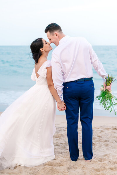 A newly wed couple rubbing noses while walking the beaches of Wilmington holding hands by JoLynn Photography, a Raleigh wedding photographer