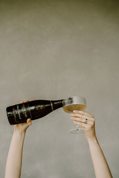 The Lovers Elopement Co - hands hold champagne glass being filled by bottle in the air