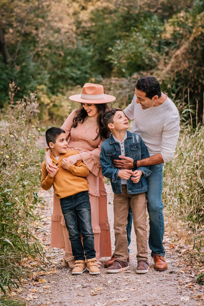 A family of four walks along a nature trail, with the parents lovingly guiding their two young sons, both of whom look up at them with wide smiles.