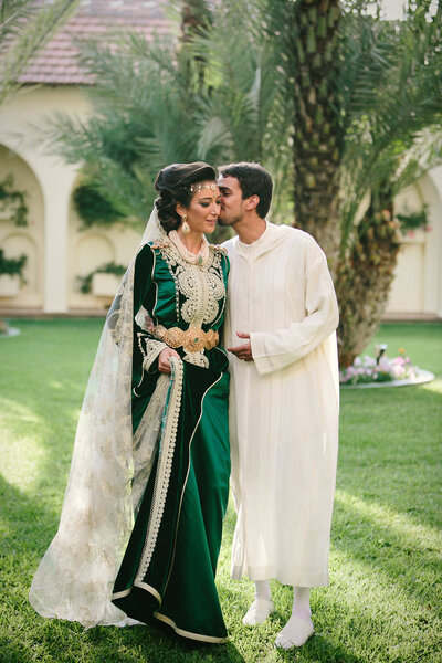Groom in traditional white attire kissing bride in a luxurious green Moroccan caftan with intricate gold embroidery and sheer veil, standing in a palm-filled garden courtyard. A romantic and culturally rich wedding portrait capturing elegance and intimacy.