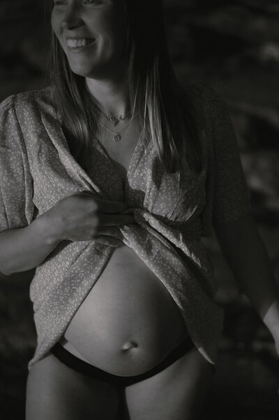 Gravid kvinna står i profil med skjorta på sig i solnedgång.