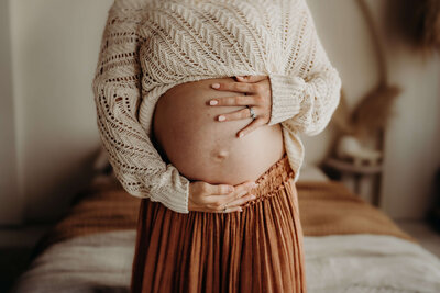 A close-up shot of a first time expectant mother, rubbing her pregnant belly while wearing a unique cream, colored crochet sweater and a brown maxi skirt with a bare belly.