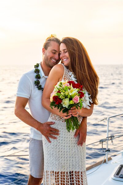 Newly engaged couple embracing on a sailboat in Hawaii after their proposal, smiling as the woman holds a bouquet with the ocean behind them.