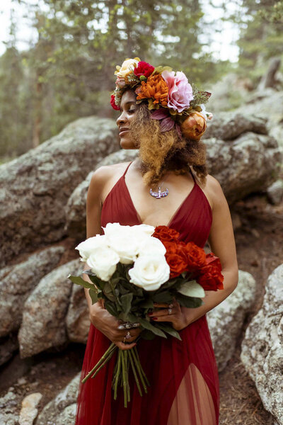 woman with a flower crown and a red dress standing in the forest with a red and white rose bouquet