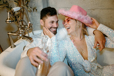 Bride in a pink hat and glasses grabbing the grooms face and both laughing. 