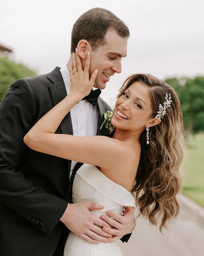 A beautiful wedding at The Royal Oaks Country Club wedding venue in Dallas, TX. A joyful bride and groom embrace, smiling warmly at each other. The bride, in an off-shoulder gown with wavy hair and a sparkling hairpiece, gently holds the groom’s face. He wears a black tuxedo and gazes at her lovingly against a soft outdoor backdrop.