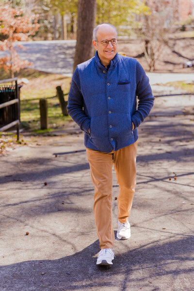 Ahmet Bozer walking hands in pockets through an Atlanta Buckhead park wearing a sleeveless jacket, beige pants, and walking shoes