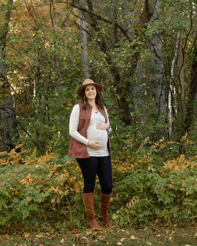 Pregnant woman with a brown hat standing among trees while smiling holding her baby bump.